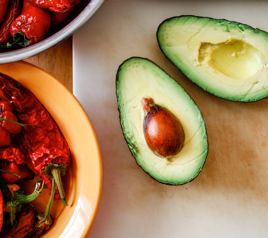 Peppers and an avocado sit on a table, ready for cooking