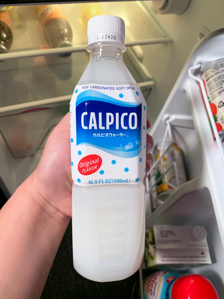 Calpico drink held by a hand in front of a refrigerator