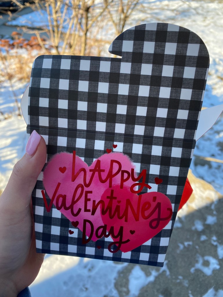 takeout container shaped box with the words happy Valentine's Day on the front. The box is held by a hand with pink nail polish in front of snow